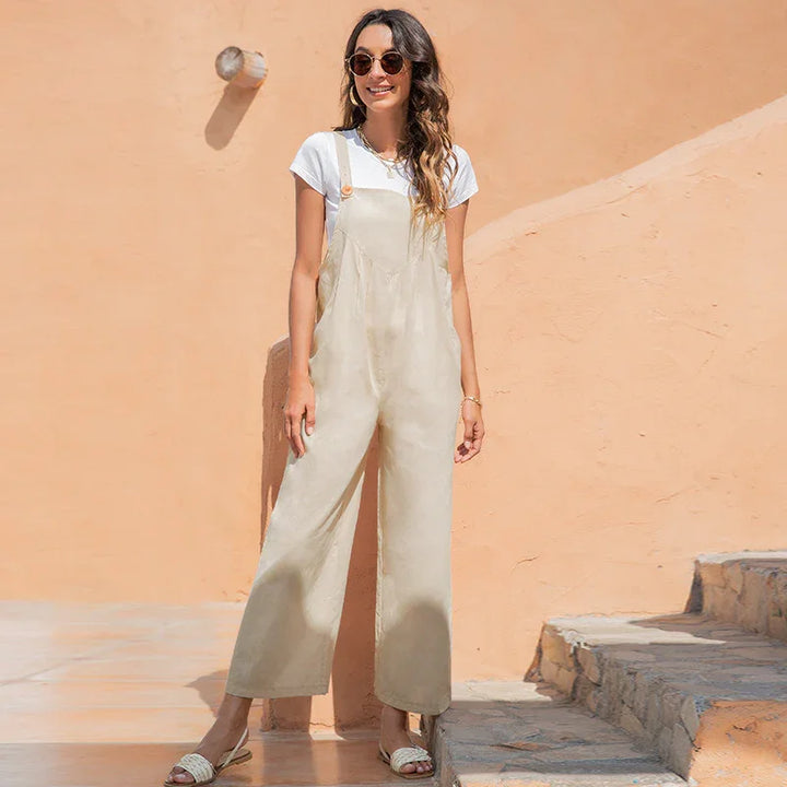 woman wearing beige wide-leg overalls, white t-shirt, sunglasses, and sandals standing by peach wall and stone steps