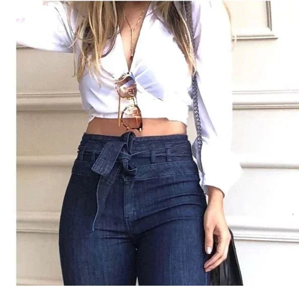 Woman wearing white crop top with knot and high-waisted dark blue jeans with belt, holding black bag