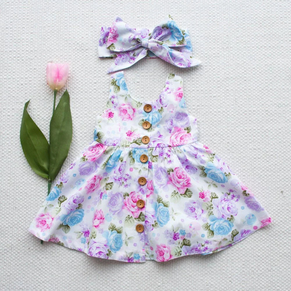 Floral baby dress with wooden buttons and matching bow headband on white textured background with pink tulip