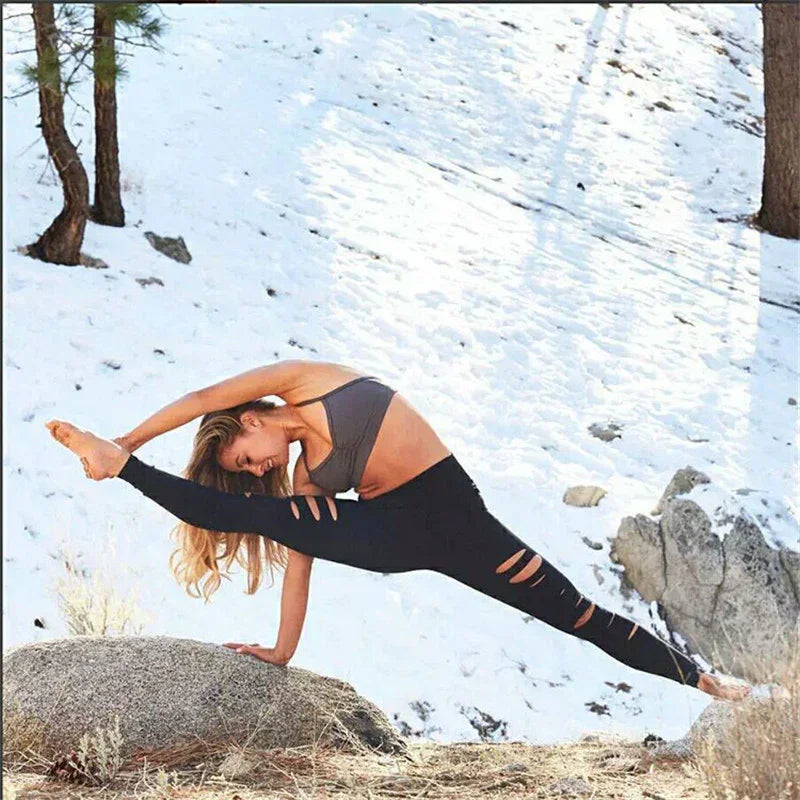 Woman in gray sports bra and ripped black leggings doing yoga side plank split on rock in snowy mountain forest