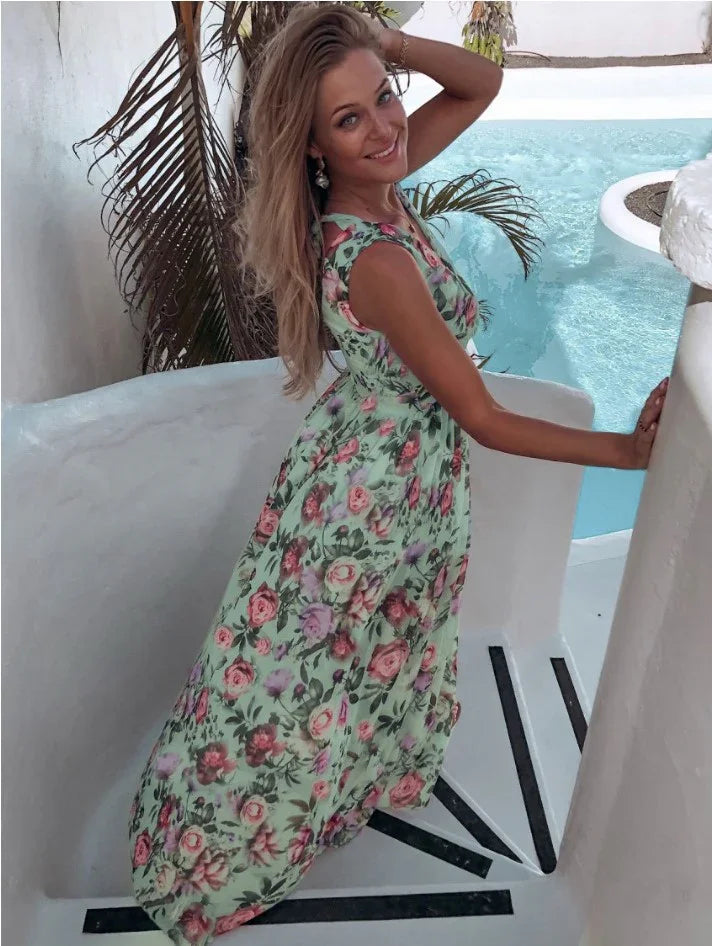 Smiling woman in a floral maxi dress posing on white stairs near a pool with tropical plants