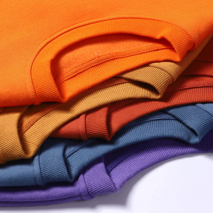 Stack of colorful folded sweatshirts in orange, brown, red, blue, and purple showing ribbed cuffs and soft fabric texture