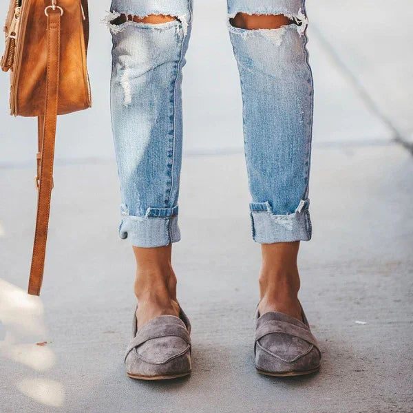 Person wearing rolled-up ripped light blue jeans and gray suede slip-on loafers on sidewalk