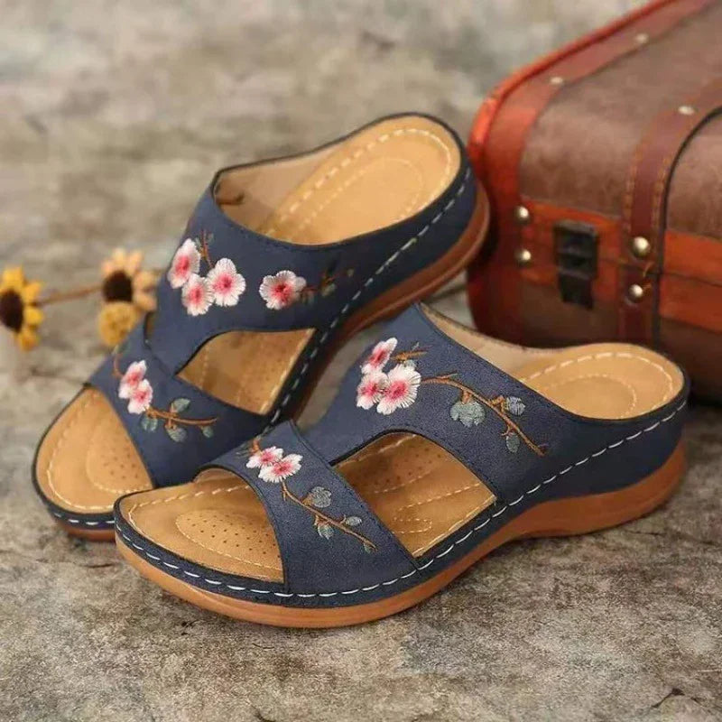 Blue slip-on sandals with pink floral embroidery and cushioned tan insoles on textured surface