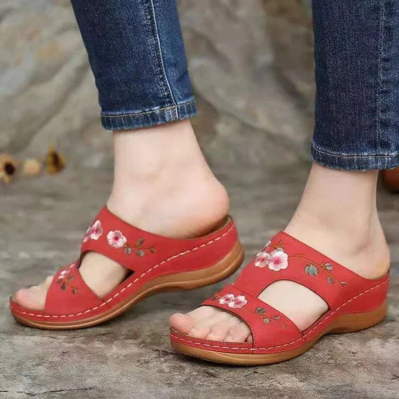 Red leather slip-on sandals with floral embroidery worn with rolled-up blue jeans on stone floor