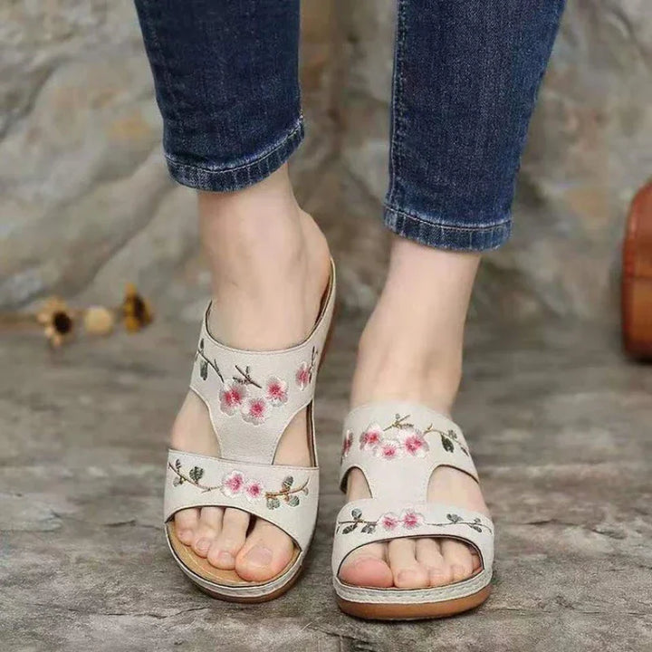 Woman wearing white floral embroidered open-toe sandals paired with blue jeans on stone floor