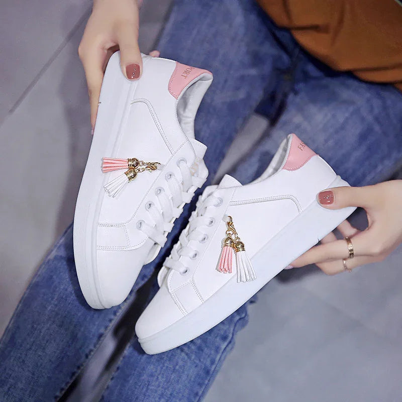 White casual sneakers with pink heel accents and decorative tassels, held by hands over blue jeans