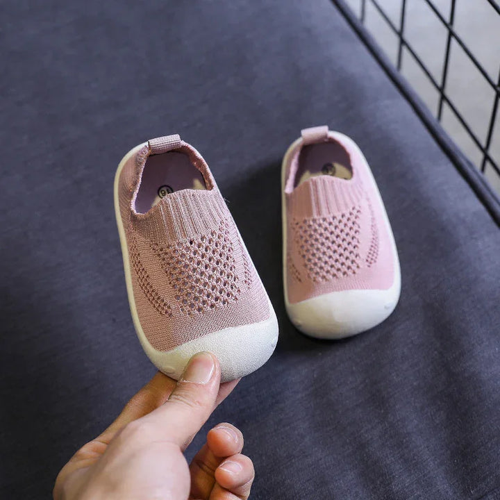 Hand holding pink baby shoes with white soles on dark fabric near black metal grid