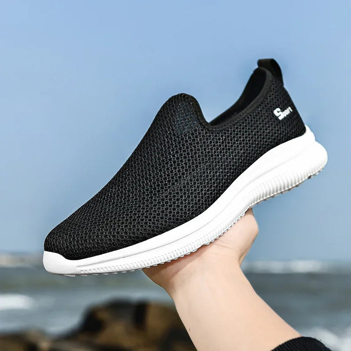 Hand holding black mesh slip-on sneaker with white sole against blue sky and ocean background