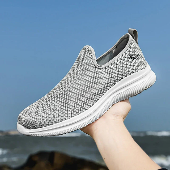 Light gray breathable mesh slip-on sneaker with white sole held against blue sky and ocean