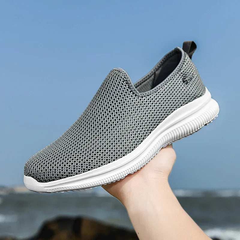 Lightweight gray mesh slip-on sneaker with white sole held outdoors by ocean shore