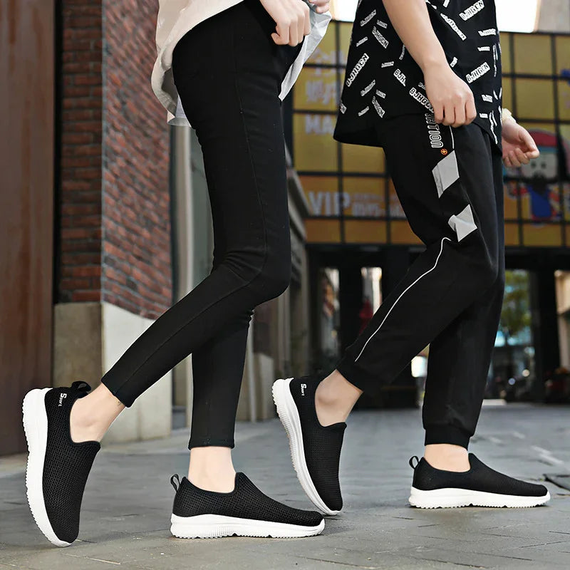Two people wearing black slip-on mesh sneakers with white soles, walking on a city sidewalk