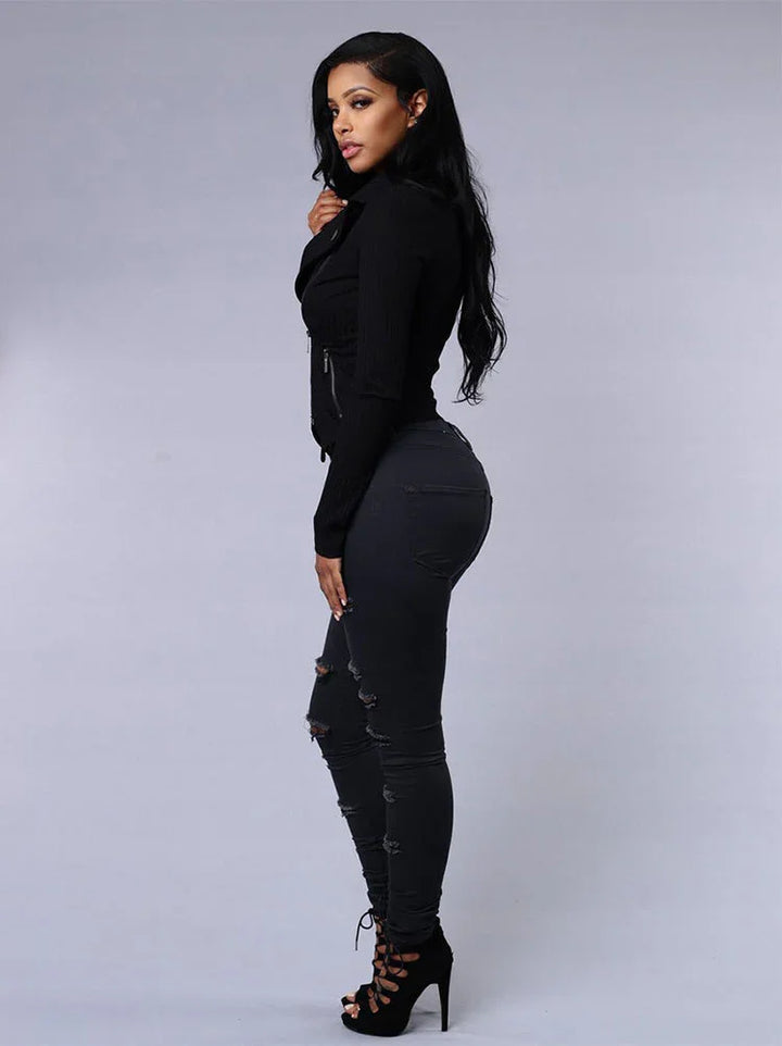 Woman in black ripped skinny jeans, black long-sleeve top, and high-heeled lace-up shoes posing against gray background
