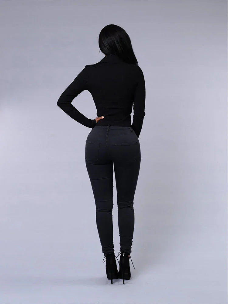 Woman wearing black fitted turtleneck, skinny jeans, and high-heeled ankle boots, standing against gray background