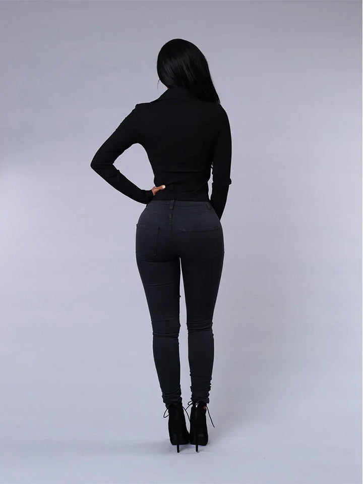 Woman wearing black fitted turtleneck, skinny jeans, and high-heeled ankle boots, standing against gray background