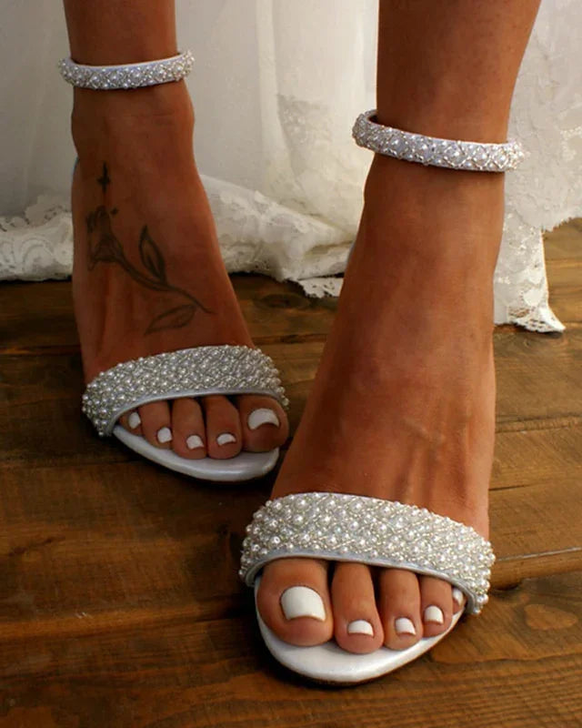 Close-up of feet wearing white pearl-embellished open-toe heels on wooden floor with white lace dress