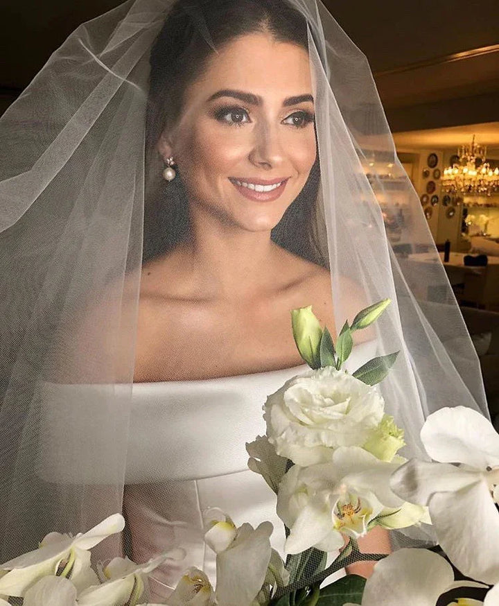 Smiling bride with pearl earrings and veil holding white flowers in elegant indoor setting