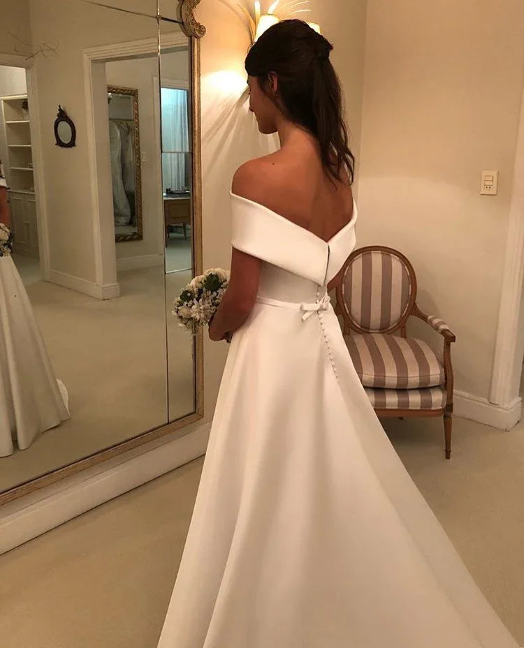 Woman in elegant off-shoulder white wedding dress holding bouquet, standing by mirror in dressing room