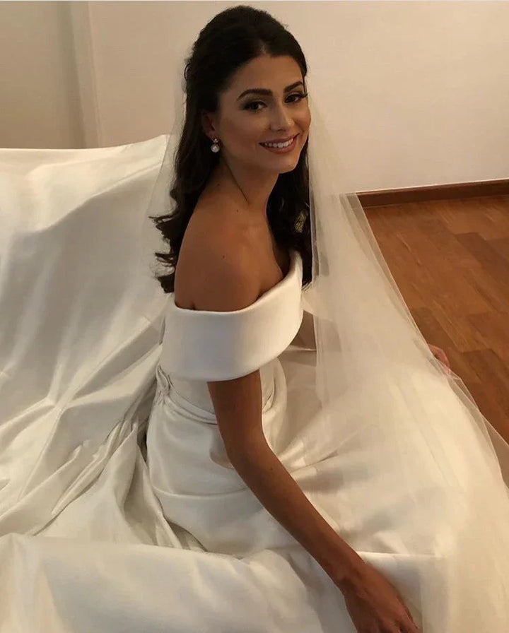 Smiling bride in an elegant off-shoulder white wedding dress with long veil sitting on wooden floor