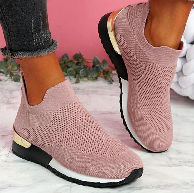 Close-up of pink knit slip-on sneakers with black soles and gold heel accents worn with frayed hem jeans on marble floor