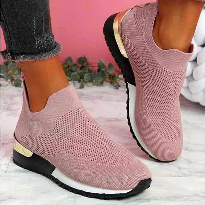 Close-up of pink knit slip-on sneakers with black soles and gold heel accents worn with frayed hem jeans on marble floor