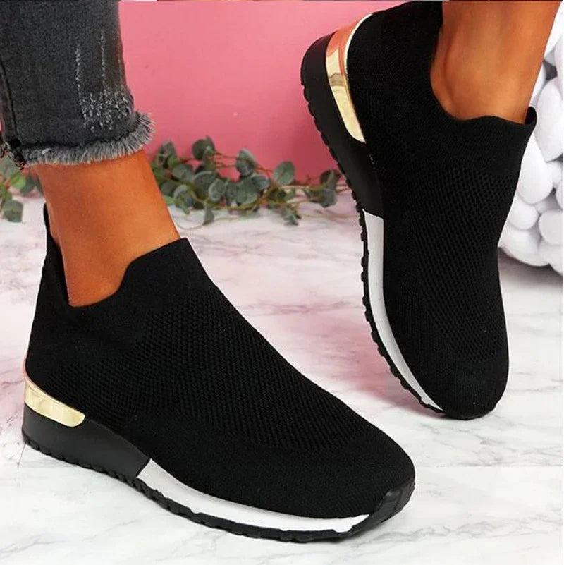Black slip-on knit sneakers with white sole and gold heel detail worn with frayed hem black jeans on marble floor