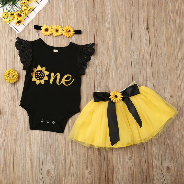Black baby bodysuit with gold sunflower design and yellow tutu skirt with black bow on wooden floor