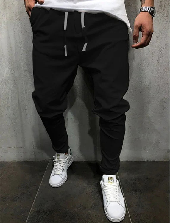 Man wearing black tapered jogger pants with white drawstrings and white sneakers against a gray wall