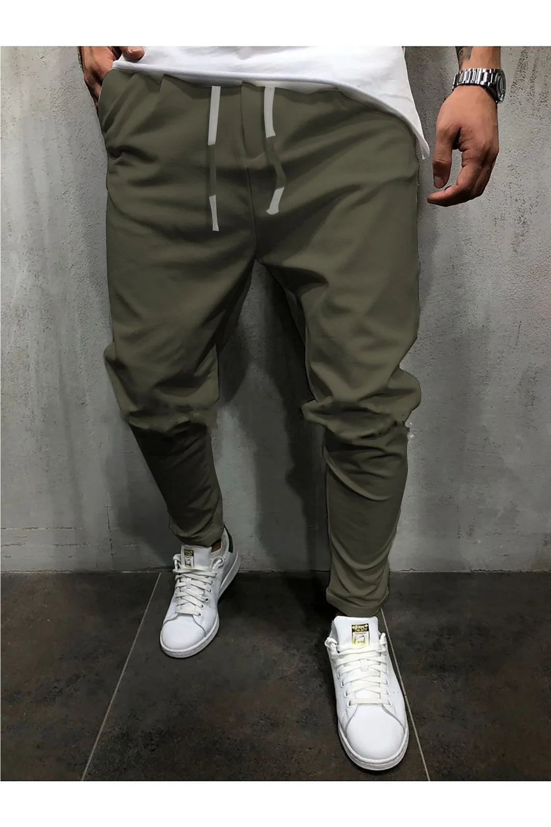 Man wearing olive green jogger pants with drawstrings and white sneakers standing against a gray wall