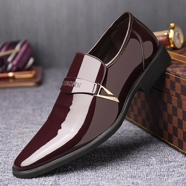Shiny burgundy leather men's dress shoe with gold buckle, displayed on gray carpet with luxury accessories