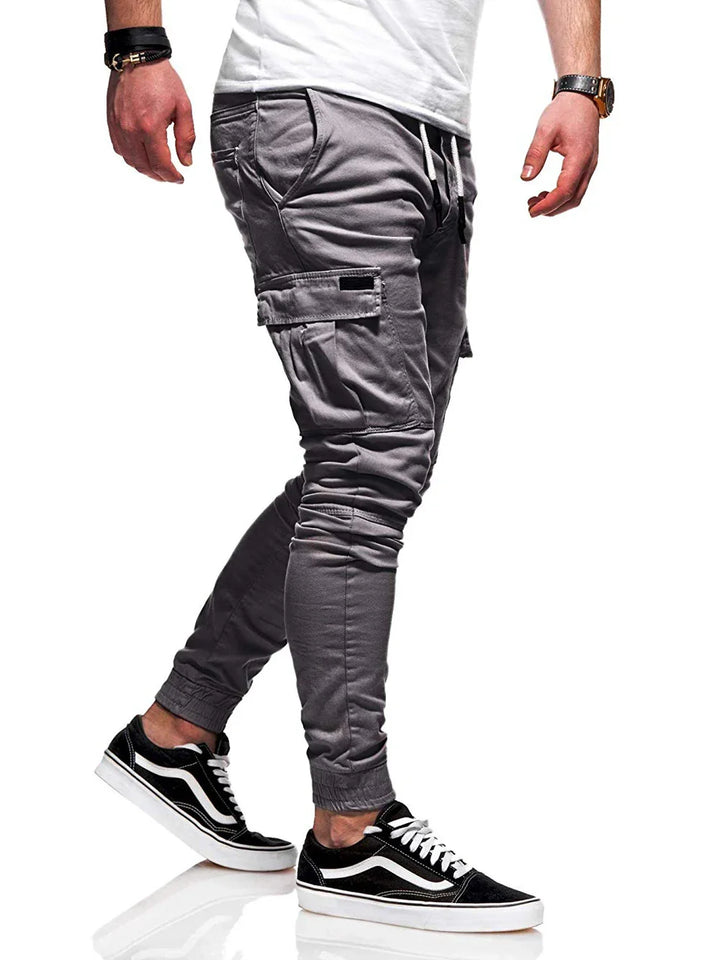 Man wearing grey cargo jogger pants with side pockets and black white sneakers on white background