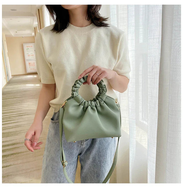 Woman wearing cream sweater and jeans holding light green ruched handle handbag in bright room
