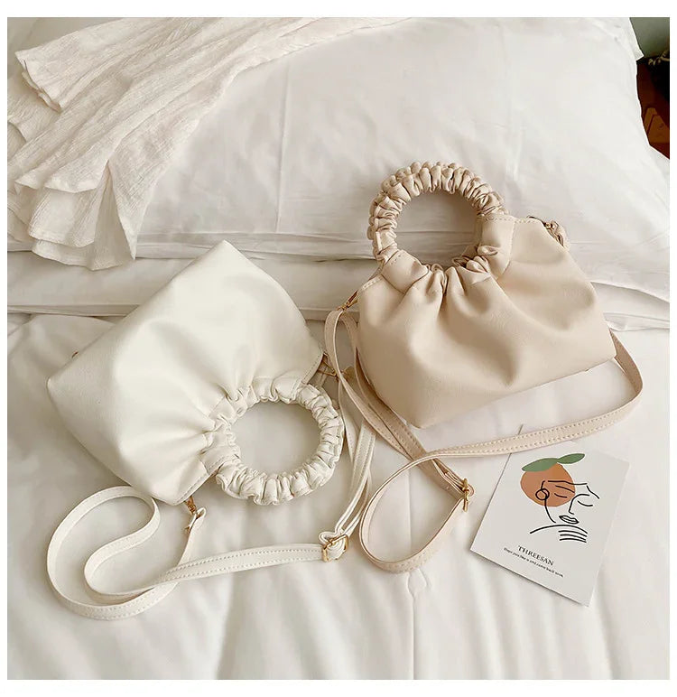 Two stylish ruched circle handle handbags in beige and cream on white bed linen with art card