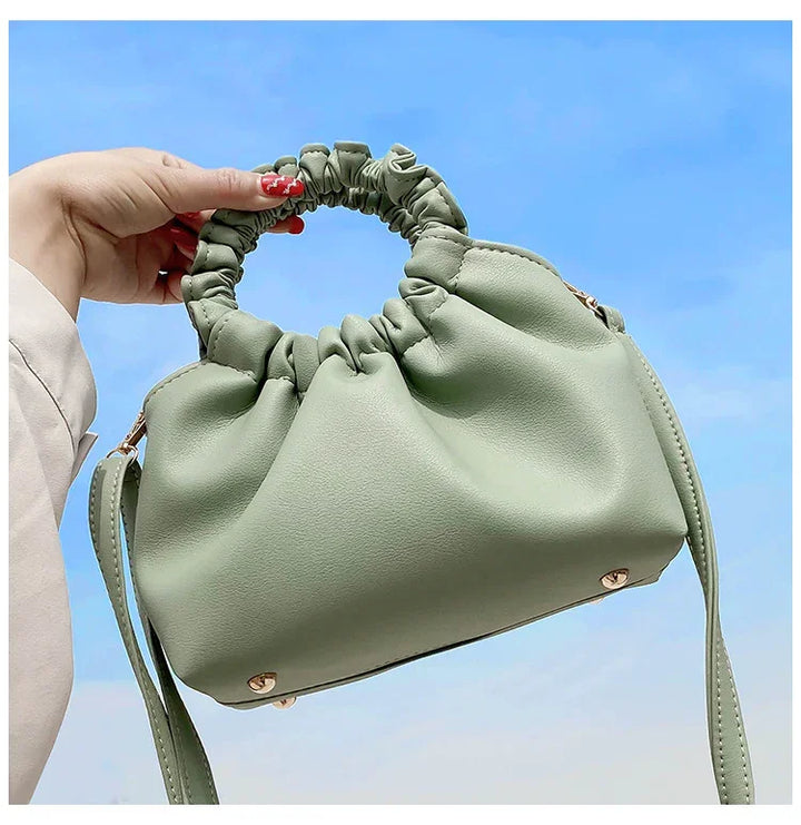 Hand holding stylish light green ruched leather handbag with round scrunchie handle against clear blue sky