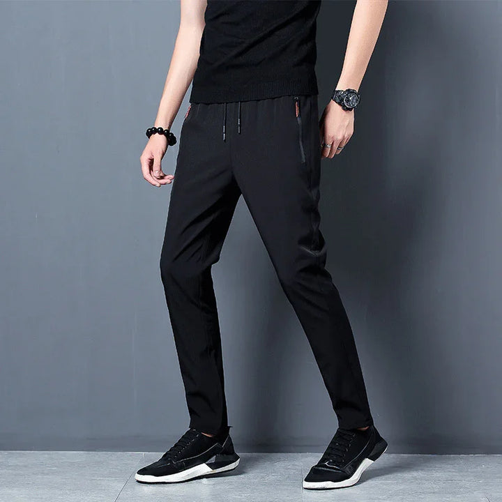 Man wearing black slim-fit casual pants, black sneakers, black top, and wristwatch against gray background