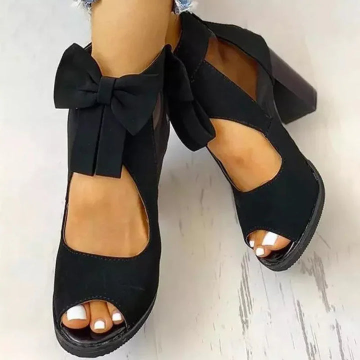Black open-toe block heels with bow detail on light wood floor, worn with white pedicure