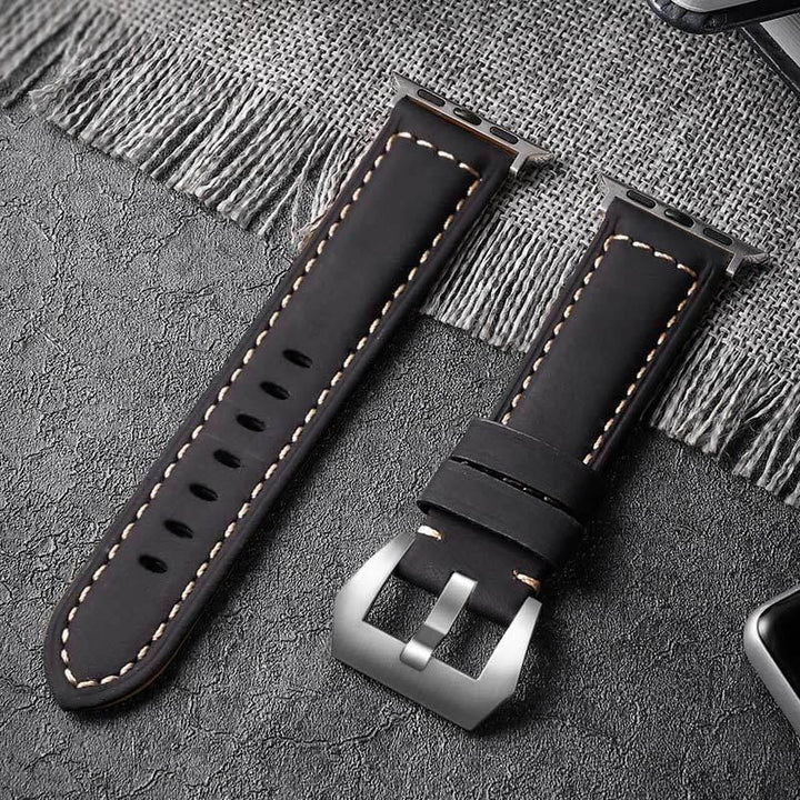 Black leather watch strap with beige stitching and silver buckle on textured gray surface