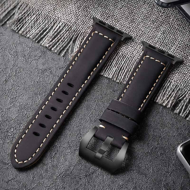 Black leather watch band with beige stitching and matte black buckle on textured surface