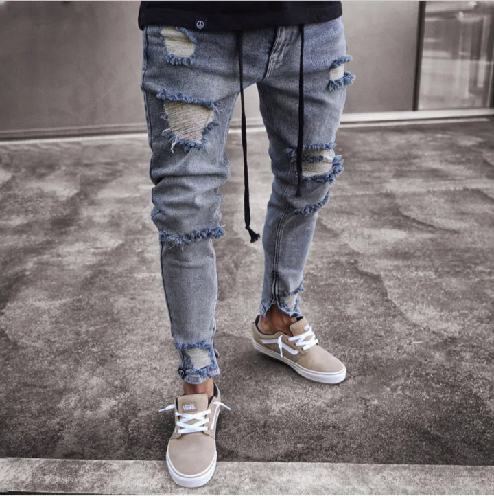Slim fit distressed blue jeans with frayed patches worn with beige Vans sneakers on concrete floor