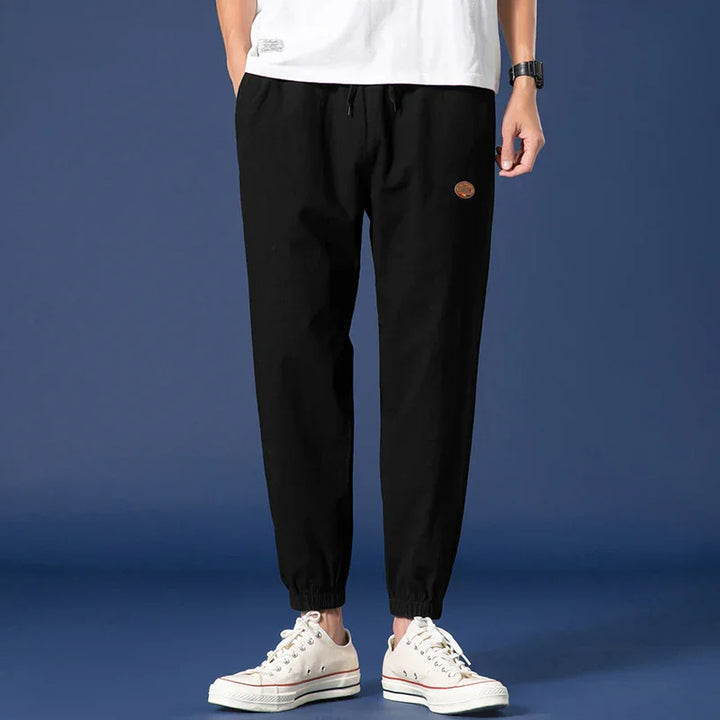 Man wearing black casual jogger pants with elastic cuffs and white sneakers on blue background