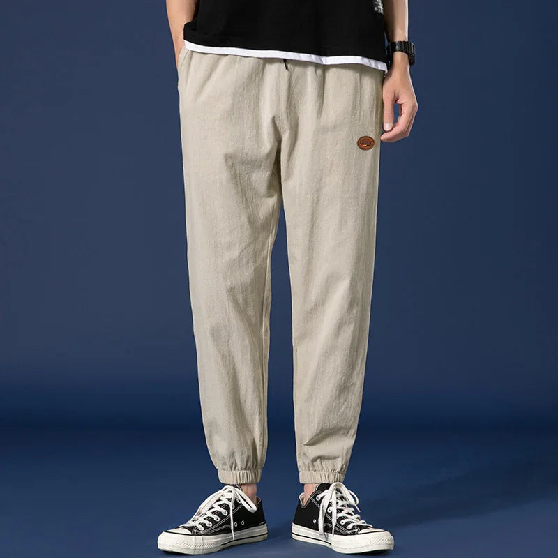 Man wearing beige casual jogger pants with elastic cuffs and black sneakers against dark background