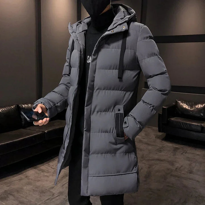 Man wearing gray puffer winter coat with hood, standing indoors holding a smartphone