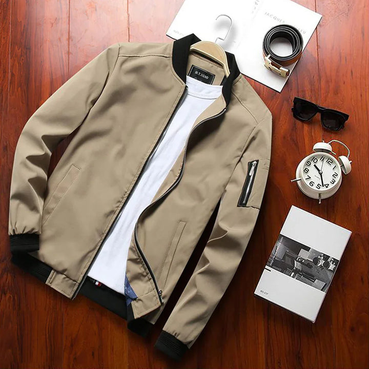 Beige bomber jacket with black collar and cuffs displayed on wooden floor with white t-shirt, black sunglasses, alarm clock, belt, and book