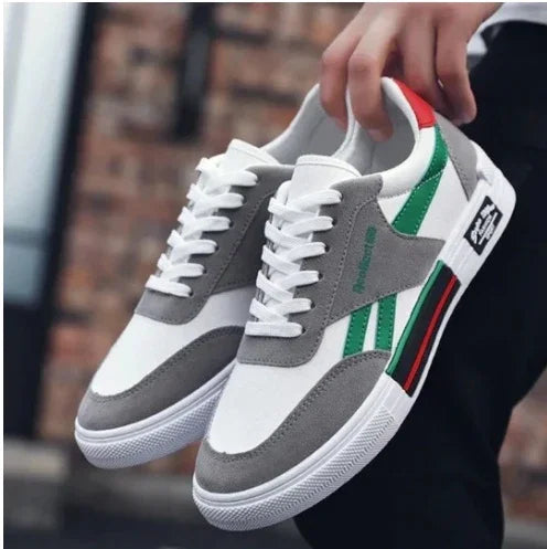 Casual white and gray sneakers with green and red stripe accents, held by hand against blurred brick wall