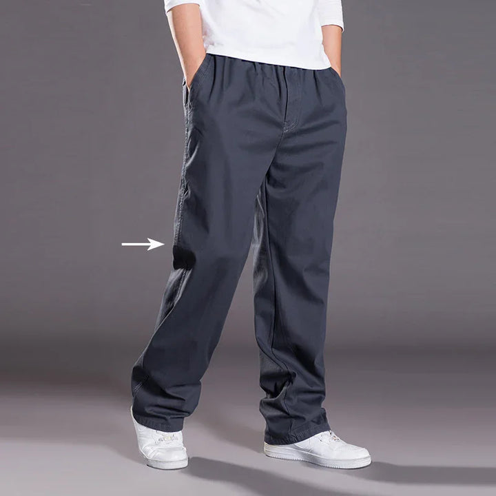 Person wearing dark gray casual pants with hands in pockets and white sneakers on gray background