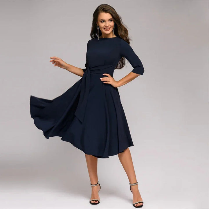 Smiling woman wearing a navy blue midi dress with 3/4 sleeves and black high heels on gray background