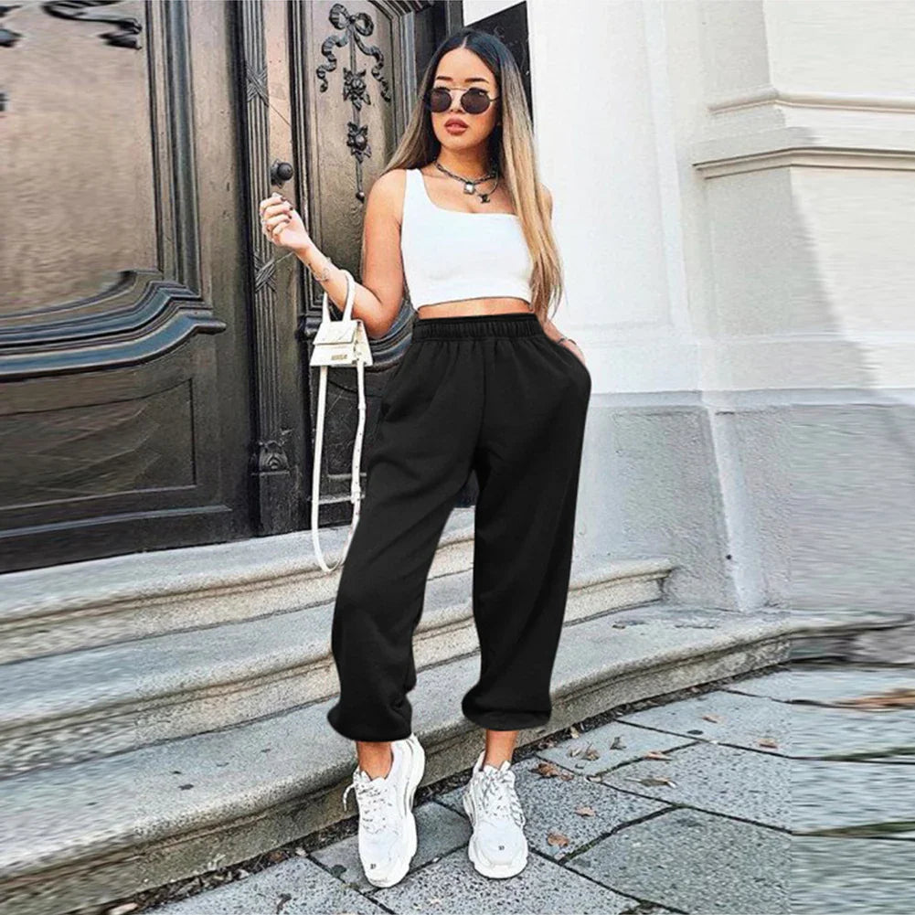 Woman wearing black jogger pants, white crop top, white sneakers, and sunglasses standing on outdoor steps