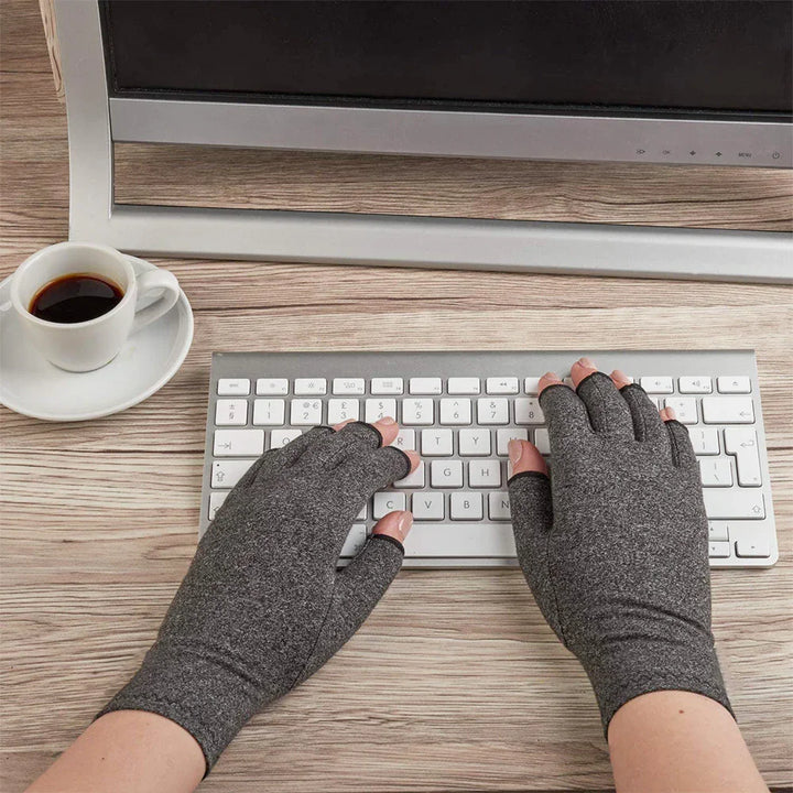Hands wearing gray fingerless compression gloves typing on white keyboard with coffee cup on wooden desk