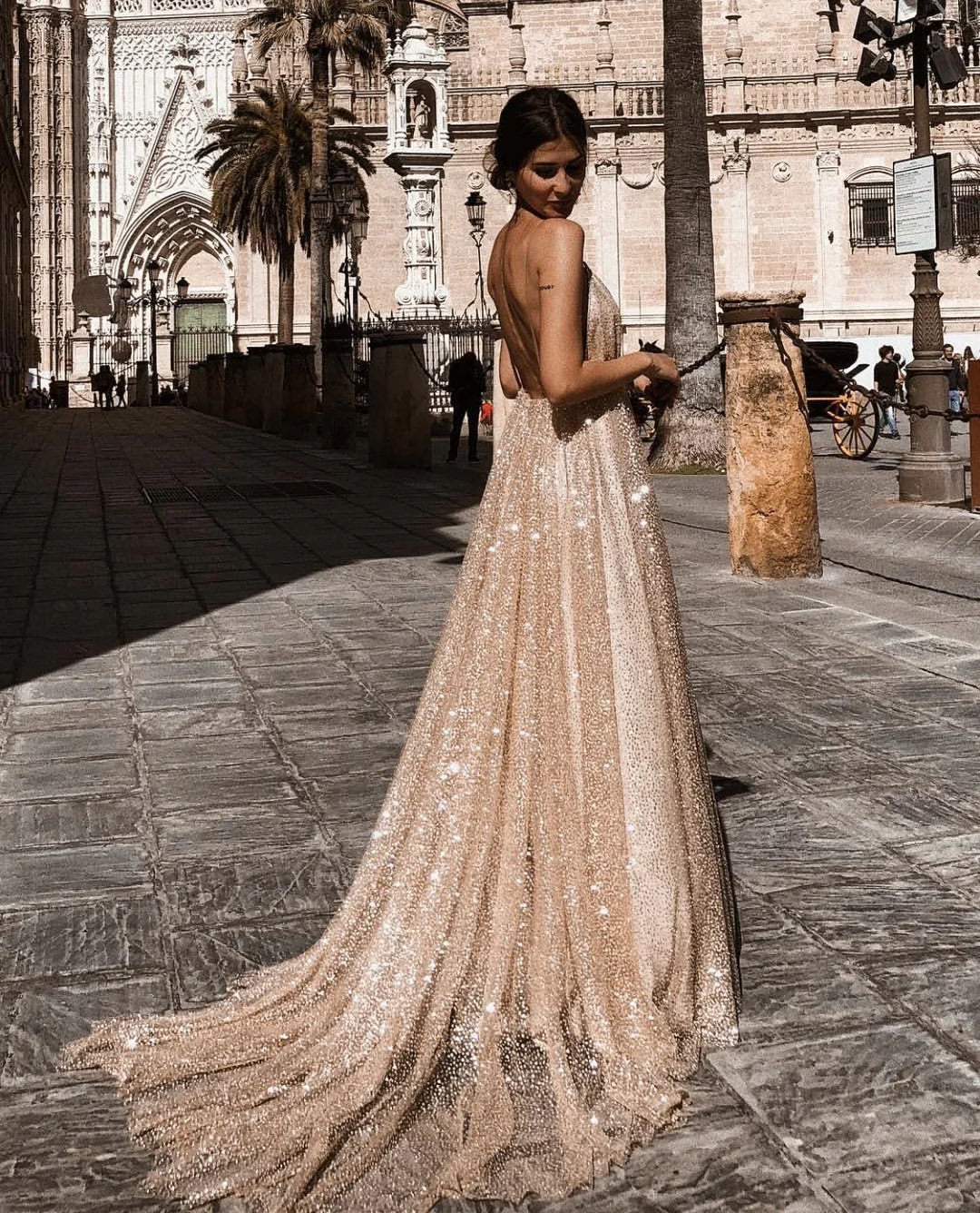 Woman wearing a sparkling backless beige gown standing on a historic stone street with gothic architecture