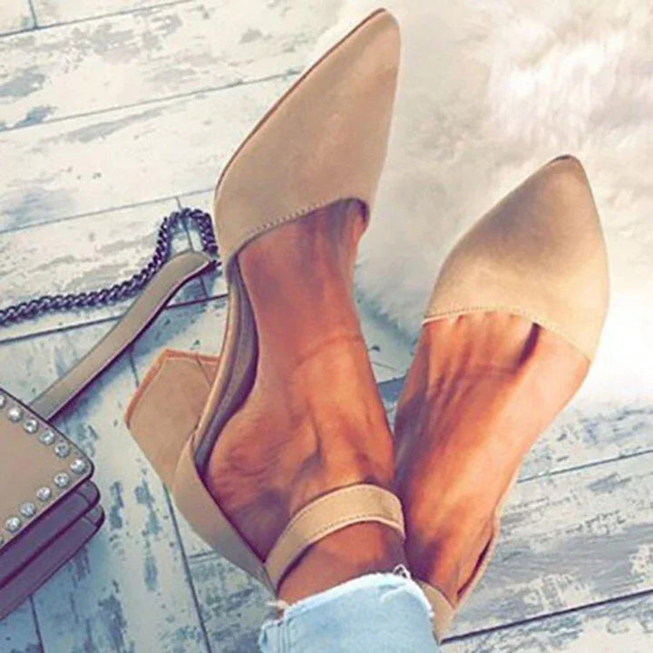 Beige pointed toe flats with ankle strap worn on feet over distressed gray wooden floor near studded handbag
